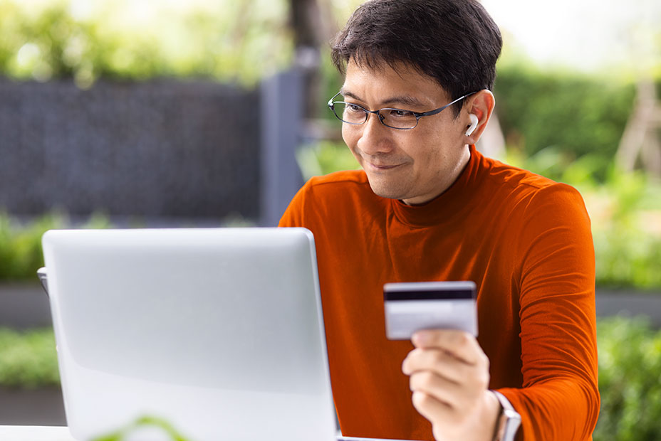 A smiling person wearing glasses looking at a laptop while holding a credit card.
